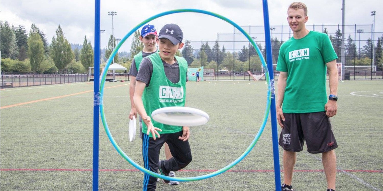 A child with autism throws a flying disc through a hoop under the guidance of a male volunteer.