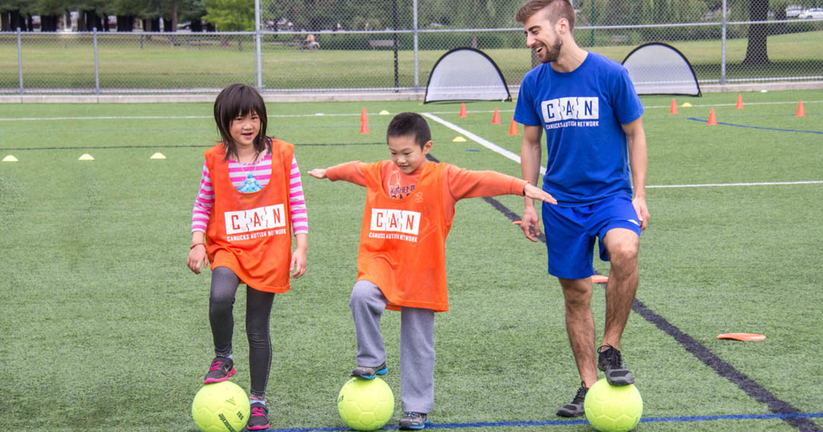 A female child, a male child and an adult male stand in a row on an outdoor turf field, each with a soccer ball under one of their feet.