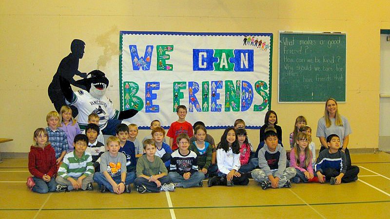 An elementary school classroom sits in front of a "We CAN Be Friends" sign with an orca whale mascot seated behind them in a school gym.