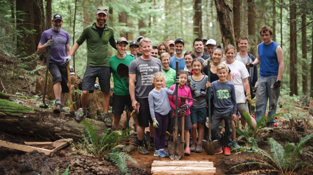 A group photo of adults and kids with shovels on a mountain trail.